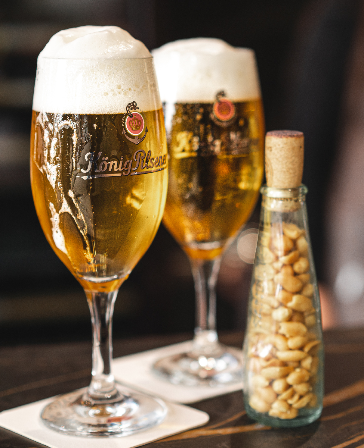 material-03 Zwei Gläser König Pilsener mit Schaumkrone, daneben eine Glasflasche mit Erdnüssen.
